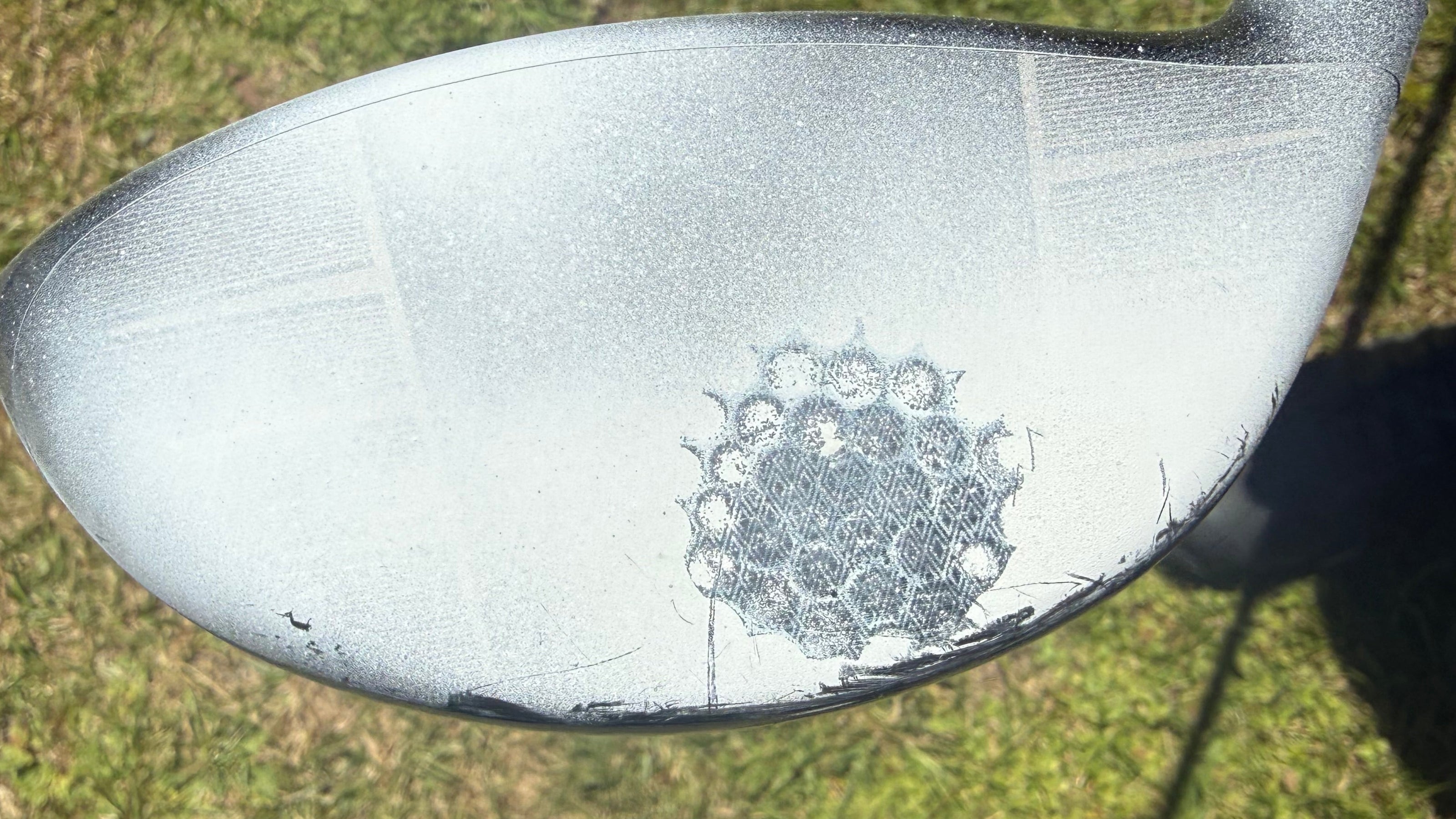 A close-up of a golf club head on grass, showing a circular pattern of golf balls embedded in the clubface. A person's leg and shoe are partially visible on the right side.